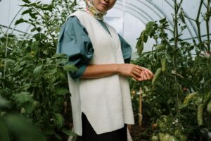 A woman gardener in stylish attire tending to lush tomato plants inside a greenhouse.