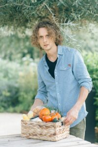 Caucasian man holds a basket of fresh produce outdoors, surrounded by greenery.